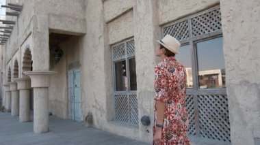 Dubai 'de Al Seef Parkı' nı gezen etnik giysili ve şapkalı genç bir kadın. Geleneksel Emirati mimarisi ve modern atmosferiyle çevrelenmiş olan sanatçı, Dubai Dereleri kültür mirasının cazibesine sahip..