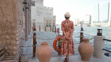 Dubai 'de Al Seef Parkı' nı gezen etnik giysili ve şapkalı genç bir kadın. Geleneksel Emirati mimarisi ve modern atmosferiyle çevrelenmiş olan sanatçı, Dubai Dereleri kültür mirasının cazibesine sahip..