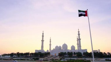 Şeyh Zayed Büyük Camii yakınlarında bayrak sallanması birlik ve mirası simgeliyor. Görkemli beyaz kubbeler ve karmaşık detaylar Emirati gururunu yansıtan sakin ve ikonik bir atmosfer yaratır..
