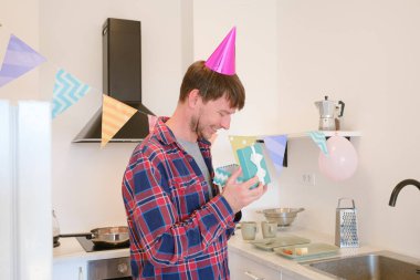 Genç adam evde hediyesini alır, şaşkınlık ve mutluluğu ifade eder. Özel bir olayı kutlarken, sıcacık, keyifli bir ortama sarılmış, neşeli ve unutulmaz bir anı yakalayan hediyeye hayran kalır.