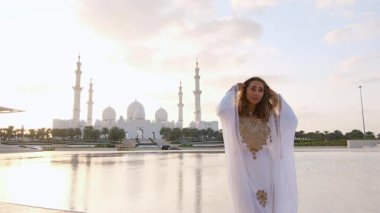 Abu Dabi 'deki Şeyh Zayed Büyük Camii yakınlarındaki Wahat Al Karama' daki geleneksel Arap elbiseli kadın, gün batımında kültürel zarafet ve ikonik İslami mimari sergiliyor..