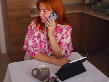 Genç bir kadın ev mutfağında tablet üzerinde çalışıyor. Gerçek bir serbest çalışan kadın evden uzakta çalışmak için bilgisayar kullanır. akıllı telefon ve kadın kontrol uygulaması, web sitesi veya sosyal medya