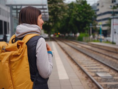Frankfurt 'ta tramvay durağında duran turist kadın, şehir merkezinin ortasında, şehir hayatı ve modern binalarla çevrili bir şekilde toplu taşıma için bekliyor ve şehrin günlük ritmini yaşıyor..