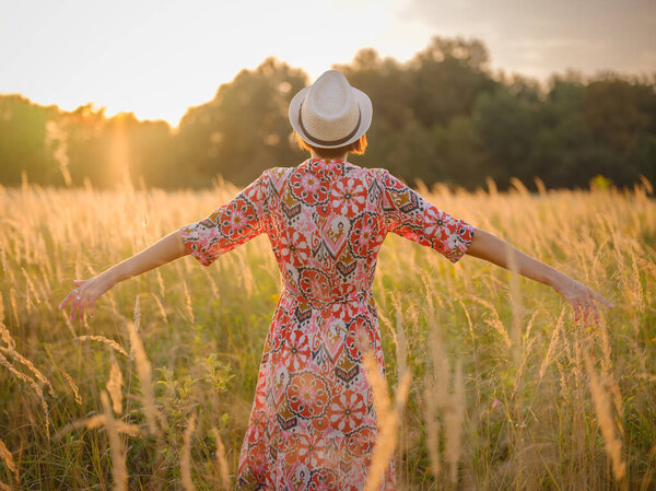 Молодая женщина, идущая по живописному европейскому полю в конце лета. Золотой солнечный свет, пышная зелень и спокойная сельская атмосфера создают мирную сельскую местность.