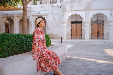 Dubai 'de Al Seef Parkı' nı gezen etnik kıyafetli ve şapkalı genç bir kadın. Geleneksel Emirati mimarisi ve modern atmosferiyle çevrelenmiş olan sanatçı, Dubai Dereleri kültür mirasının cazibesinden hoşlanıyor..