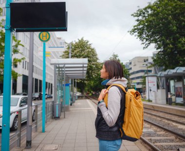 Frankfurt 'ta tramvay durağında duran turist kadın, şehir merkezinin ortasında, şehir hayatı ve modern binalarla çevrili bir şekilde toplu taşıma için bekliyor ve şehrin günlük ritmini yaşıyor..