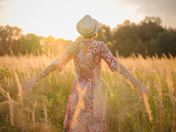 Молодая женщина, идущая по живописному европейскому полю в конце лета. Золотой солнечный свет, пышная зелень и спокойная сельская атмосфера создают мирную сельскую местность.