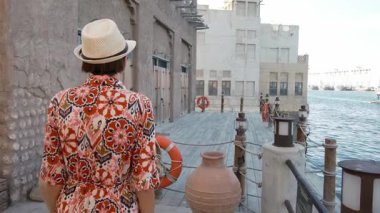 Dubai 'de Al Seef Parkı' nı gezen etnik giysili ve şapkalı genç bir kadın. Geleneksel Emirati mimarisi ve modern atmosferiyle çevrelenmiş olan sanatçı, Dubai Dereleri kültür mirasının cazibesine sahip..