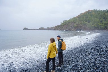 Çiftler Türk kıyı şeridini seviyor. Taşlarda oturmak, deniz dalgalarını izlemek, birlikte dinlenmek, sakin mavi suyun yanında, yumuşak kış ışığı altında romantik bir an..