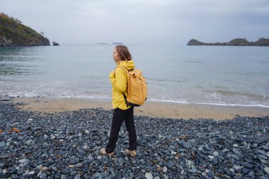 Bulutlu kış denizinin yakınında yürüyen bir kadın. Taşlarda oturmak, deniz dalgalarını izlemek, birlikte dinlenmek, sakin mavi suyun yanında, yumuşak kış ışığı altında romantik bir an..