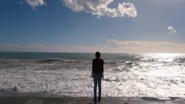 Türkiye 'nin Antalya kentinde fırtınalı Akdeniz kıyıları yakınında duran genç bir kadın. Güneşli kış günü, güçlü dalgalar, rüzgarlı saçlar, derin düşünceler. Doğayla barışçıl ama dramatik bir bağ anı..