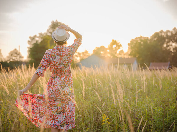 Молодая женщина, идущая по живописному европейскому полю в конце лета. Золотой солнечный свет, пышная зелень и спокойная сельская атмосфера создают мирную сельскую местность.