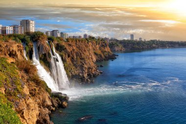Picturesque morning landscape of the Lower Duden waterfall against the backdrop of the steep shores of the Mediterranean Sea and urban development in the Antalya city, Turkey                               