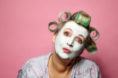 Komik, düşünceli, saçları yuvarlanan ve pembe arka planda kilden maske takan son sınıf öğrencisi bir kadın.