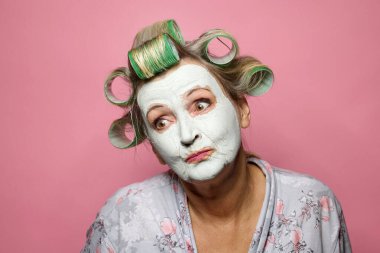 Komik, düşünceli, saçları yuvarlanan ve pembe arka planda kilden maske takan son sınıf öğrencisi bir kadın.