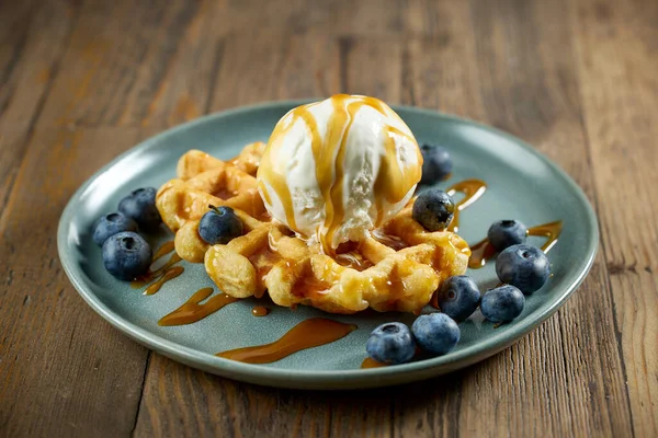 Bir tabak Belçika waffle 'ı vanilyalı dondurma, karamel sosu ve ahşap masanın üstünde yabanmersini.