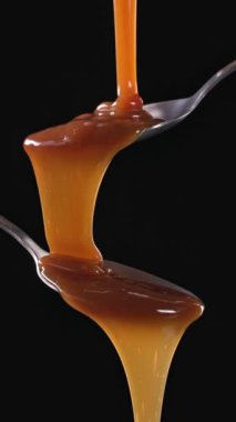 Erimiş karamel sosu siyah arka planda metal kaşıktan akıyor. Yemek yakın çekim, ağır çekim, dikey video 4K