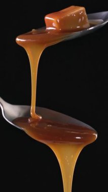 Erimiş karamel sosu siyah arka planda metal kaşıktan akıyor. Yemek yakın çekim, yavaş çekim, dikey video 4K yakınlaştır