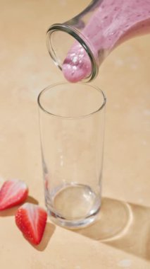 Taze yapılmış pembe çilek ve muz suyu güneşli bir masa yüzeyindeki bir şişeden bir bardağa akıyor.