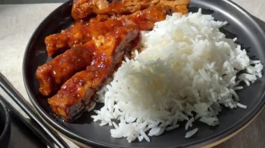 Siyah tabakta sarımsak soslu yumuşak domuz etiyle servis edilen haşlanmış beyaz pirinç porsiyonu. Kamera usulca yakınlaştırıyor, yemeğin karmaşık detaylarını vurguluyor, aşçılık için mükemmel.