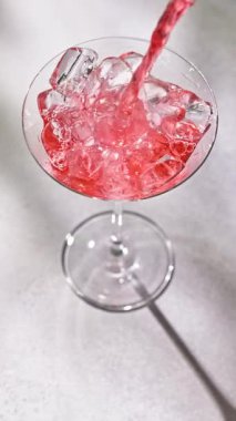 Gri, güneşli bir restoran masasında buz küpleriyle kokteyl bardağına dökülen taze kırmızı içki.