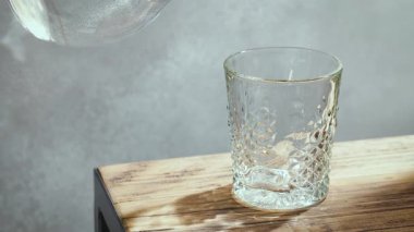 Güneşli ahşap mutfak masasında bardağa dökülen temiz su ve arka plan gri, ağır çekim.