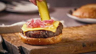 Aşçı ahşap kesim tahtasında lezzetli çizburger yapmak için ızgara burgerin üzerine salatalık dilimleri koydu.