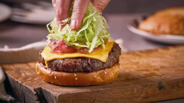 Aşçı, ahşap kesme tahtasına lezzetli çizburger yapmak için ızgara burgerin üzerine doğranmış marul koydu.