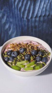 Jogurt ve meyveli beyaz sağlıklı kahvaltı kasesi mavi arka planda dönüyor, yakınlaş, yiyecek yakın plan