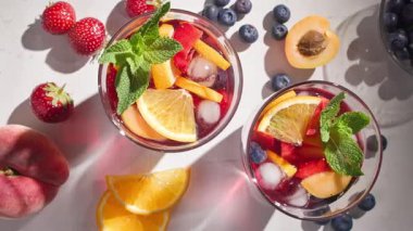 Kırmızı sangria bardaklarına yakın çekim, taze meyve parçaları, buz ve dut, nane yaprağı ile süslenmiş, üst manzara, güneşli arka plan
