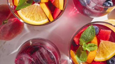 Nane yaprağıyla süslenmiş taze meyve parçaları, buz ve böğürtlen içeren dönen kırmızı sangria bardaklarının yakın plan fotoğrafı.
