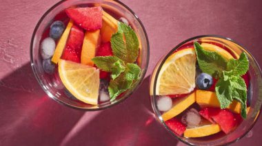 Kırmızı sangria bardaklarının yakın çekimleri taze meyve parçaları, buz ve böğürtlenlerle süslenmiş, nane yaprağı ile süslenmiş, parlak arka planda,