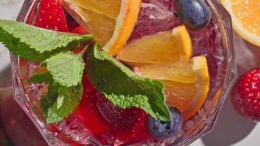 Kırmızı sangria bardağının yakın plan görüntüsü taze meyve parçaları, buz ve böğürtlen, nane yaprağı ile süslenmiş, güneşli ışık arka planında,