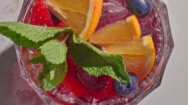 Kırmızı sangria bardağının yakın plan görüntüsü taze meyve parçaları, buz ve böğürtlen, nane yaprağı ile süslenmiş, güneşli ışık arka planında,