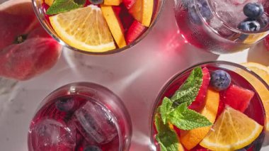 Nane yaprağıyla süslenmiş taze meyve parçaları, buz ve böğürtlen içeren dönen kırmızı sangria bardaklarının yakın plan fotoğrafı.