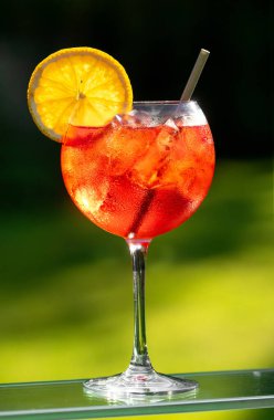 Bir bardak aperol spreyli kokteyl turuncu dilimle süslenmiş ve yeşil bahçe arka planında saman var.