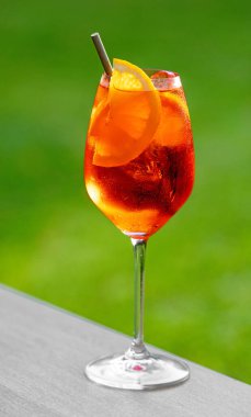 Bir bardak aperol spreyli kokteyl turuncu dilimle süslenmiş ve yeşil bahçe arka planında saman var.