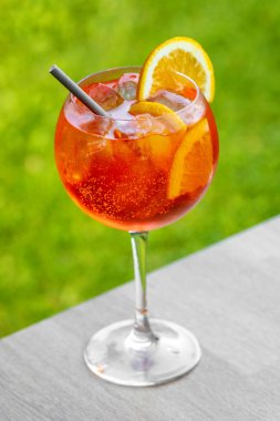 Bir bardak aperol spreyli kokteyl turuncu dilimle süslenmiş ve yeşil bahçe arka planında saman var.