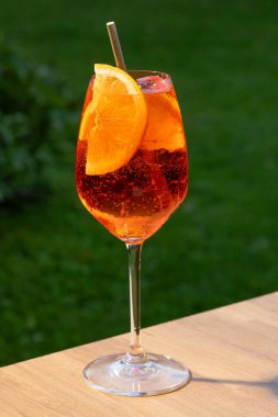 Bir bardak aperol spreyli kokteyl turuncu dilimle süslenmiş ve yeşil bahçe arka planında saman var.