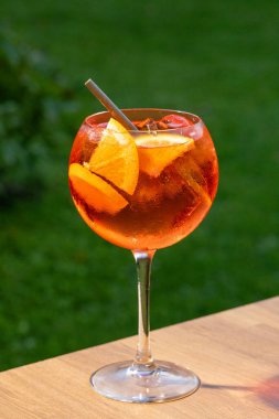Bir bardak aperol spreyli kokteyl turuncu dilimle süslenmiş ve yeşil bahçe arka planında saman var.
