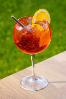 Bir bardak aperol spreyli kokteyl turuncu dilimle süslenmiş ve yeşil bahçe arka planında saman var.