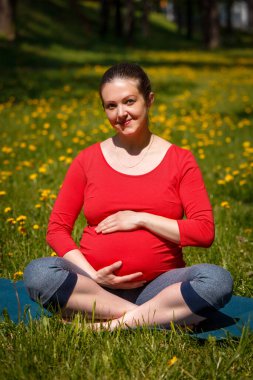 Hamilelik yogası - hamile gülümseyen kadın Asana Sukhasana 'ya gülümsüyor kolay yoga pozu veriyor yaz aylarında karahindiba ile çimlerin üzerinde karnını tutuyor.