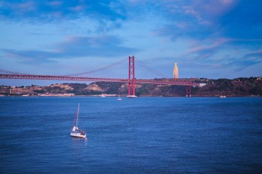 25 de Abril Köprüsü 'nün Tagus nehri üzerindeki ünlü turistik mekan, Kral İsa Anıtı ve alacakaranlıkta bir yat teknesi manzarası. Lizbon, Portekiz