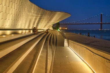 Lizbon, Portekiz - 08 Ağustos 2022: MAAT - Sanat, Mimarlık ve Teknoloji Müzesi, Tagus nehrinin kıyısında, arka planında 25 Nisan akşamı köprü bulunan modern bir binadır. Lizbon, Portekiz