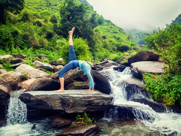 stock image Yoga outdooors - woman doing yoga asana eka pada urdva dhanurasana Upward Bow Pose back benkd outdoors at waterfall in Himalayas. Vintage retro effect filtered hipster style image.