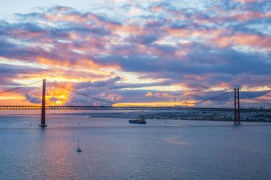 25 de Abril Köprüsü 'nün Tagus Nehri üzerinde gün batımında turistik yat tekneleri ve kargo konteynır gemisiyle Lizbon' un ünlü turistik tarihi manzarası. Lizbon, Portekiz