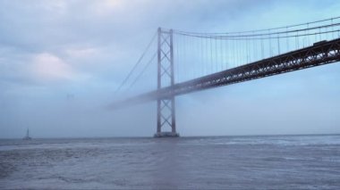 Lizbon 'un ünlü turistik beldesi 25 de Abril Köprüsü' nün Lisboa ve Almada 'yı yoğun sis bulutu altında, altından geçen teknelerle birbirine bağlayan görüntüsü. Lizbon, Portekiz