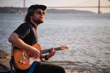 Siyahlı Hipster sokak müzisyeni Lizbon, Portekiz 'de gün batımında iskelede oturmuş elektro gitar çalıyor.