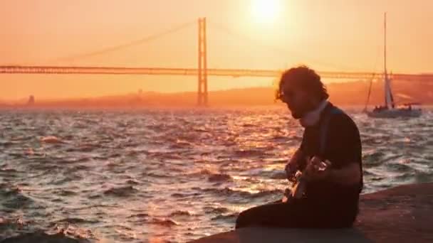 希波斯特街头音乐家穿着黑色的电吉他 在日落时分在街上的堤岸上弹奏 背景是四月二十五日的桥和游艇 葡萄牙里斯本 — 图库视频影像