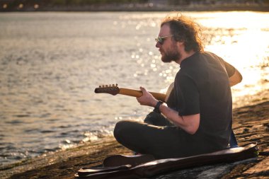 Siyahlı Hipster sokak müzisyeni gün batımında iskelede oturmuş elektro gitar çalıyor.